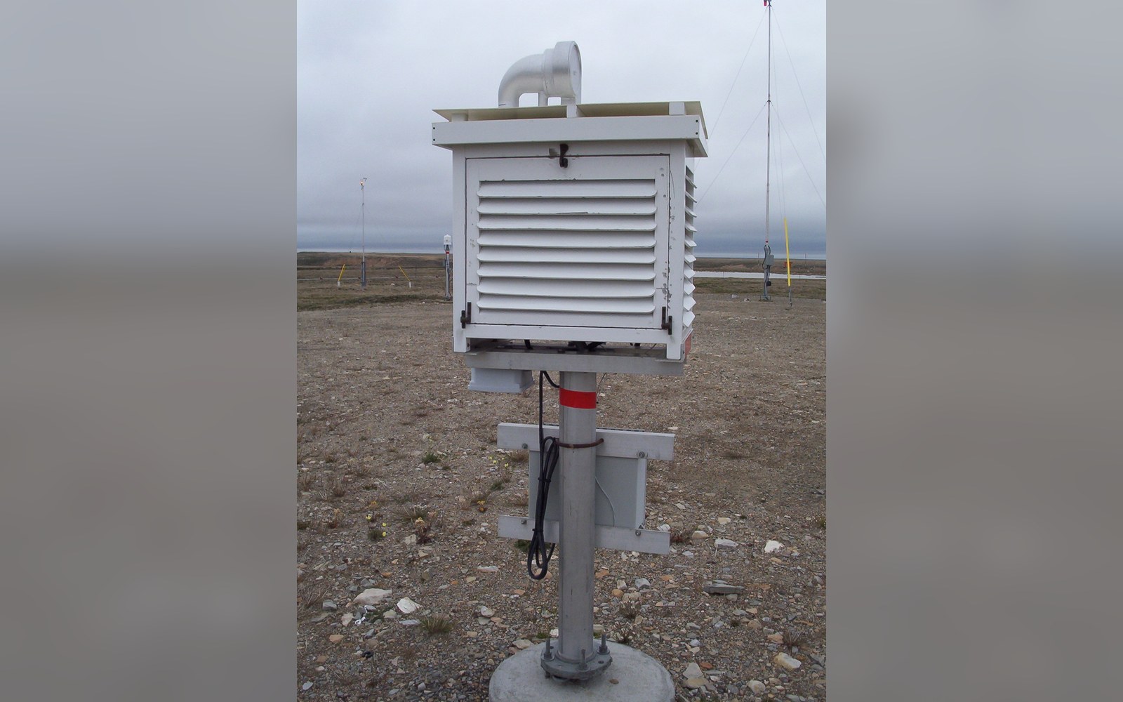 A louvered weather station enclosure mounted on a post in a barren gravel field