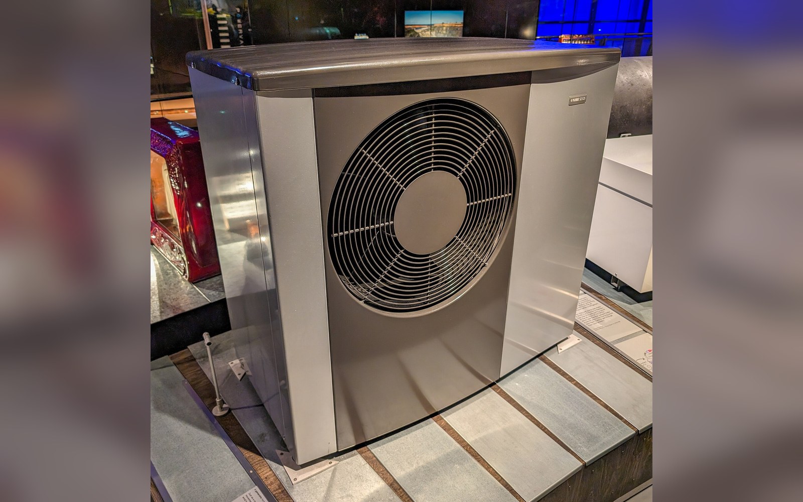 An outdoor heat pump unit with a circular fan grille on display indoors